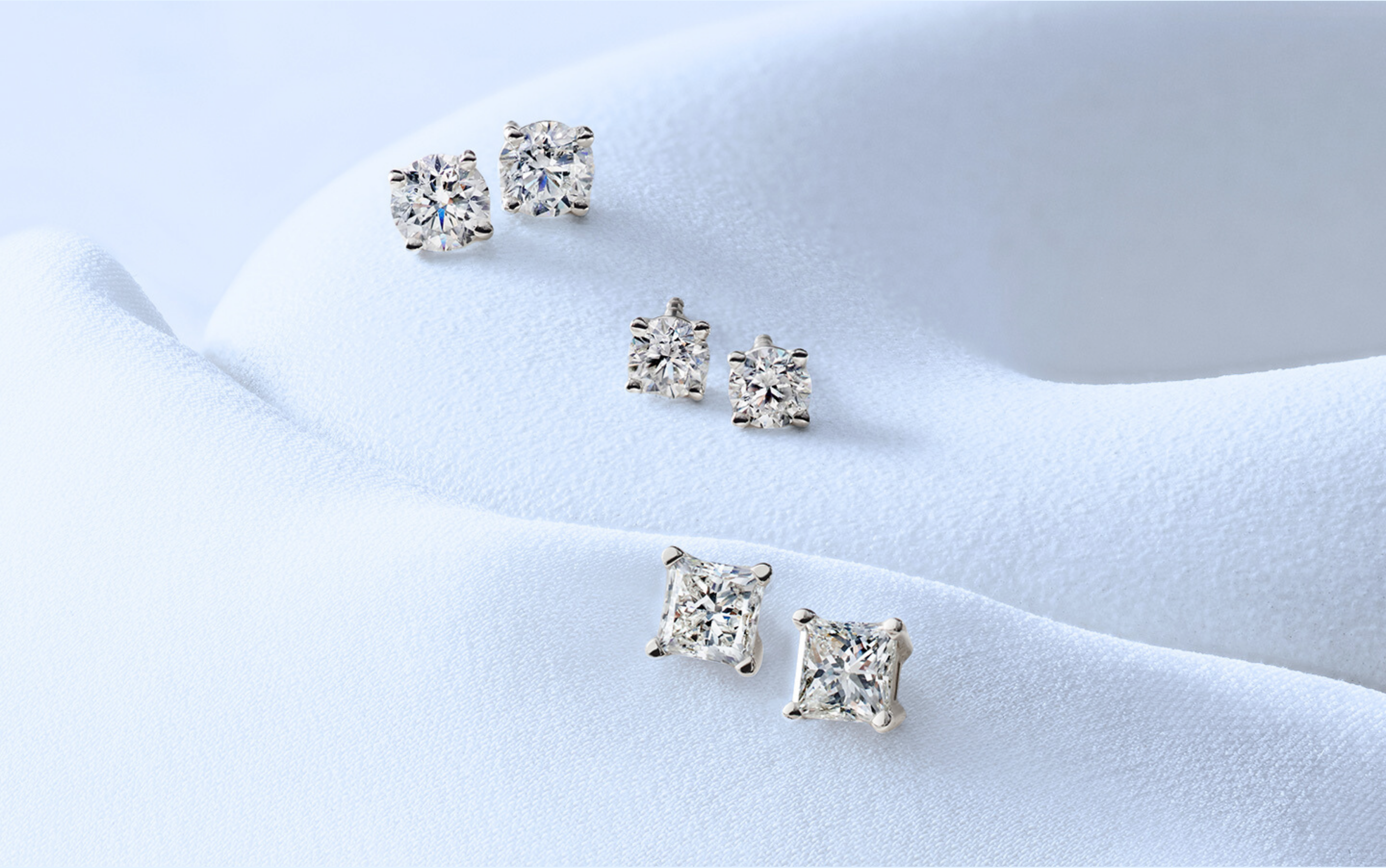 Diamond Studs Online: Elegant round and princess-cut diamond stud earrings displayed on soft white fabric.